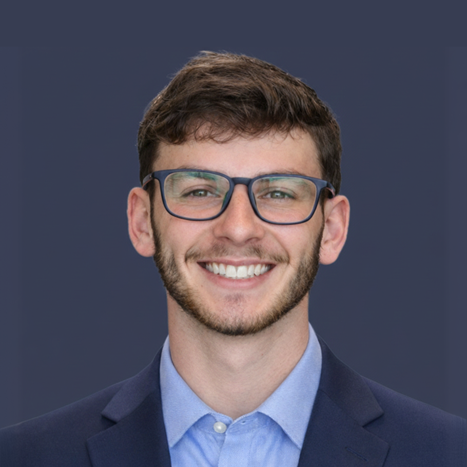Smiling man in a navy blazer and light blue shirt, wearing glasses, head-and-shoulders portrait against a slate backdrop.