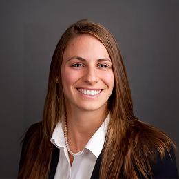 Portrait of a smiling professional woman with long brown hair, wearing a white blouse and blazer against a gray background.