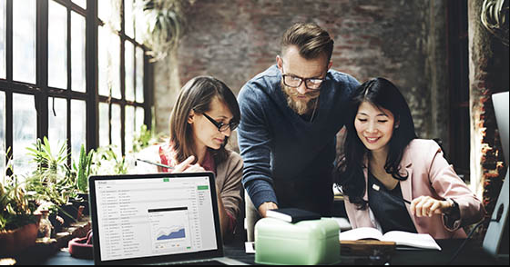 Three colleagues collaborate at a desk, reviewing a laptop graph and open notebooks in a bright office.