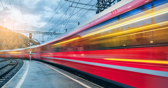Red high-speed train speeding past a platform, motion blur and overhead power lines visible.