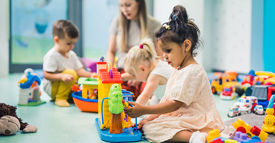 Young girl in a light dress plays with a colorful toy station on a green floor, with other children and an adult in the background.