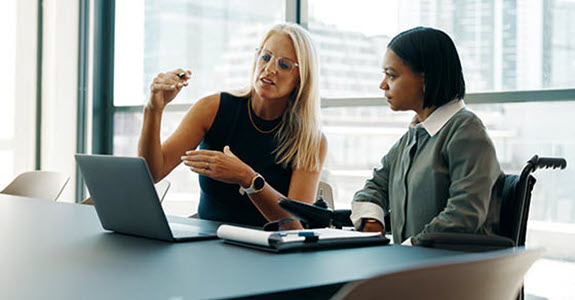 Two colleagues in a bright office discuss a presentation at a laptop, one gesturing with a pen.