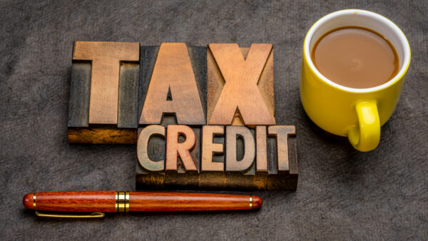 Wooden letter tiles spell 'TAX CREDIT' beside a yellow coffee mug on a dark surface; a red-brown pen lies below.