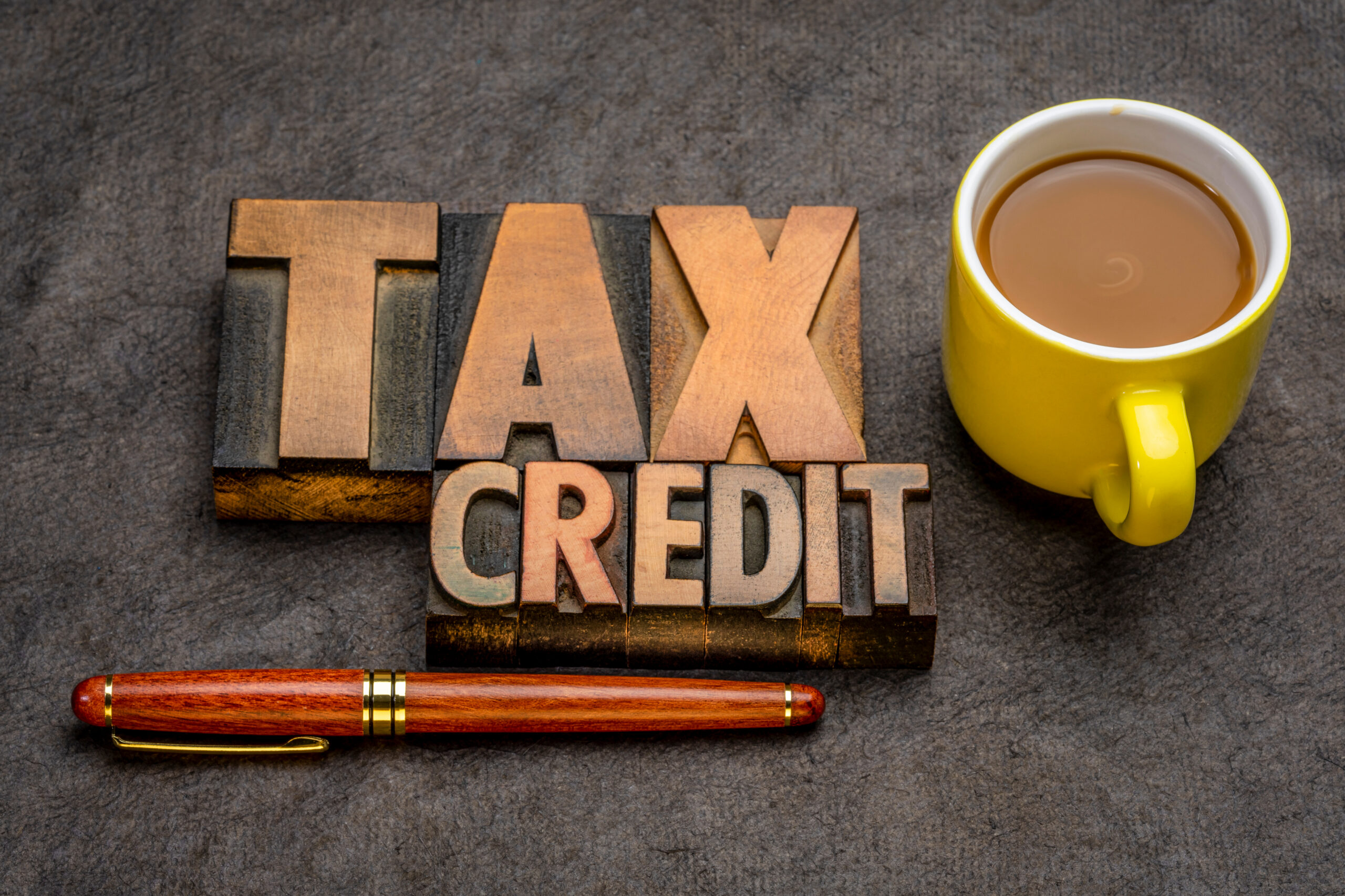 Wooden letter tiles spell 'TAX CREDIT' beside a yellow coffee mug on a dark surface; a red-brown pen lies below.
